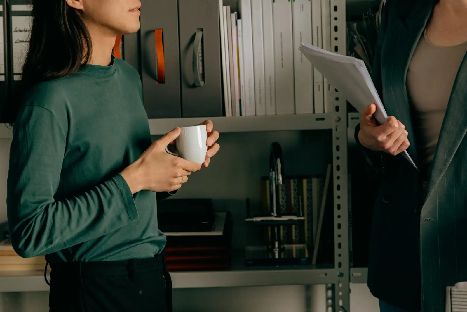 Two colleagues talking in front of a shelving unit, one holding a mug and the other a sheaf of papers